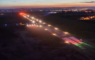 Widok z lotu ptaka na oświetlony pas startowy krośnieńskiego lotniska o zmierzchu. Wzdłuż drogi startowej świecą światła nawigacyjne, a w tle widać panoramę miasta.